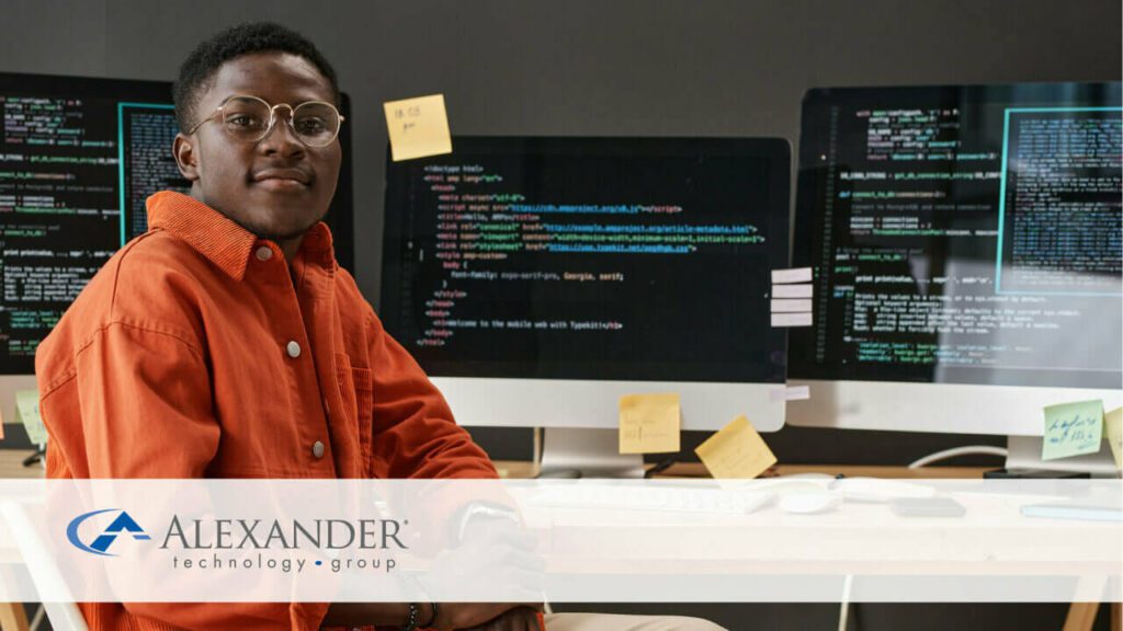a man in an orange shirt and glasses sitting in front on monitors with code on them